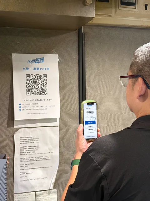 お店の壁に貼った「出勤・退勤の打刻」のQRコードを、男性がスマホで読み、打刻の画面を開いている