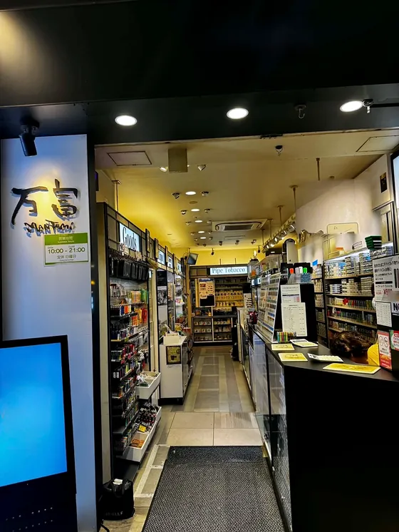 万富煙草店の入口。左に「万富」の看板、奥へ明るい通路と棚が続き、右に受付のカウンターがある