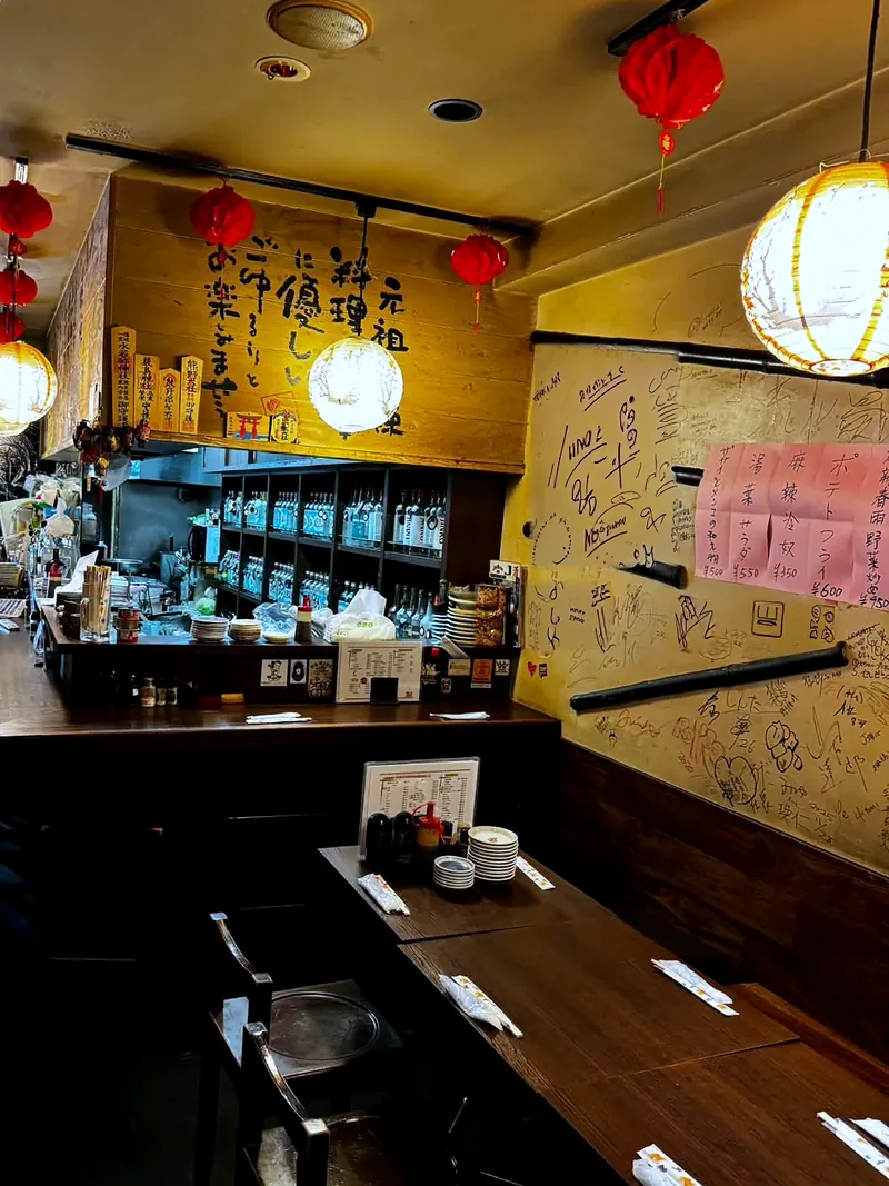 豊海屋の店内。赤いちょうちんと白いちょうちんが下がり、木の壁に大きな筆文字、寄せ書きの壁と、木のテーブル席がある