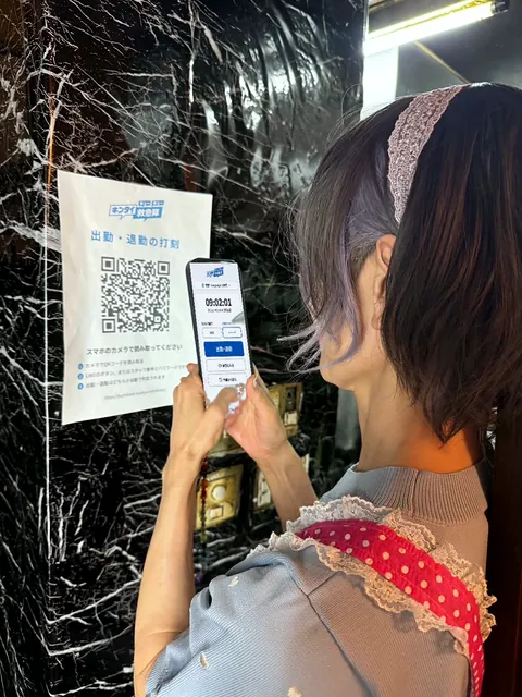 黒い大理石の壁に貼った「出勤・退勤の打刻」のQRコードを、女性がスマホで読み、打刻の画面を開いている